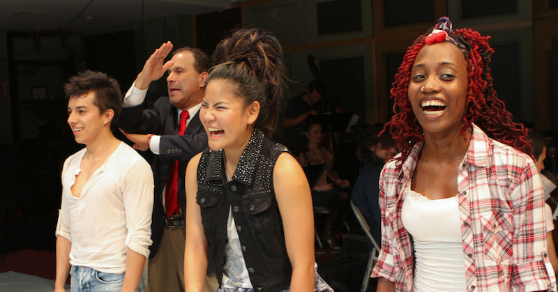 Three performers stand in a line smiling while a middle aged man in a suit executes gestures behind them